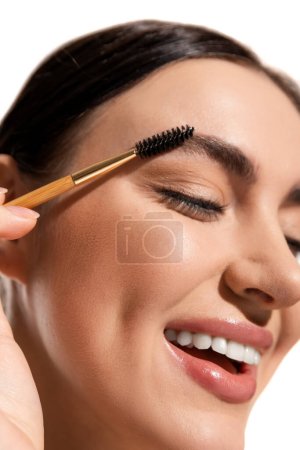 close up view of satisfied young woman styling eyebrows with brush and gel isolated on white 