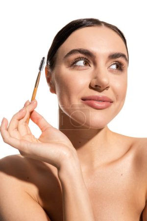 glückliche junge Frau hält Augenbrauenbürste in der Hand und schaut isoliert auf weißem Grund weg 