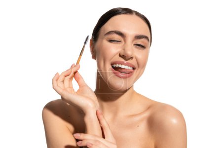 glückliche junge Frau streckt die Zunge heraus, während sie Augenbrauenbürste isoliert auf Weiß hält 