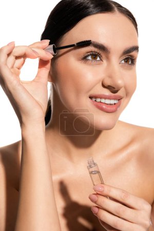 Foto de Happy and brunette woman styling eyebrows with brush and holding styling gel isolated on white - Imagen libre de derechos
