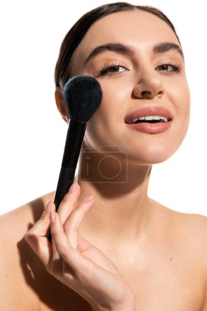 brunette woman with bare shoulders holding soft powder brush near cheek isolated on white 
