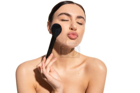 Foto de Morena mujer con hombros desnudos sosteniendo suave pincel en polvo y pucheros labios aislados en blanco - Imagen libre de derechos