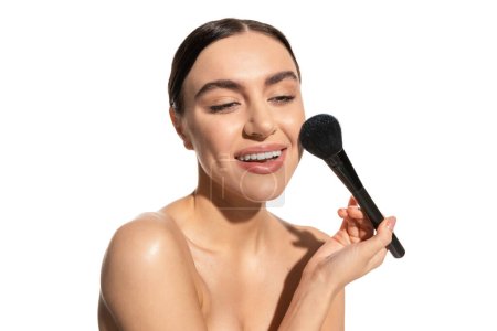 cheerful young woman with bare shoulders holding soft powder brush near cheek isolated on white 