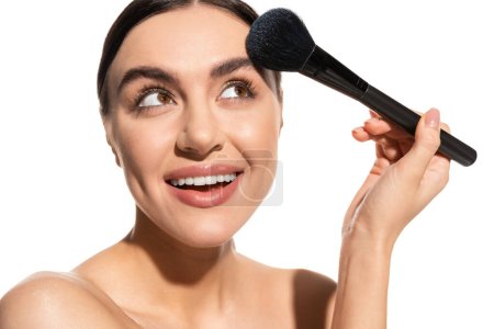 mujer sonriente con hombros desnudos sosteniendo cepillo de polvo suave cerca de la cara aislada en blanco 