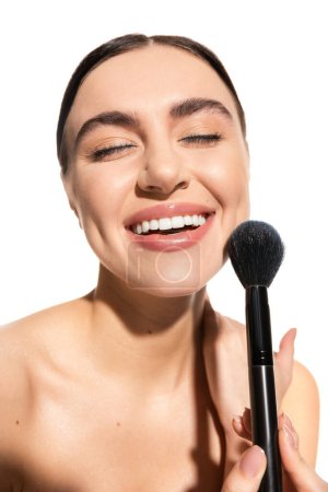 Téléchargez les photos : Satisfied woman with bare shoulders holding soft powder brush near face isolated on white - en image libre de droit