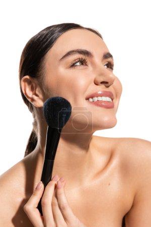 satisfied woman with bare shoulders holding soft powder brush near cheek isolated on white 