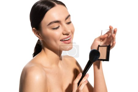 satisfied woman with bare shoulders holding soft cosmetic brush near powder blush isolated on white 