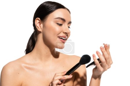 Téléchargez les photos : Cheerful woman with bare shoulders holding soft cosmetic brush near powder blush isolated on white - en image libre de droit