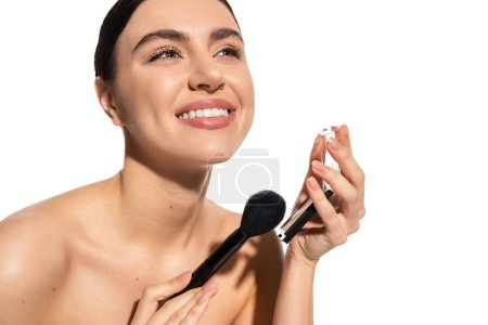 Photo for Pleased woman with bare shoulders holding cosmetic brush near powder blush isolated on white - Royalty Free Image