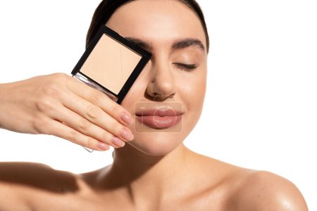 young woman covering eye while holding neutral beige face powder isolated on white 