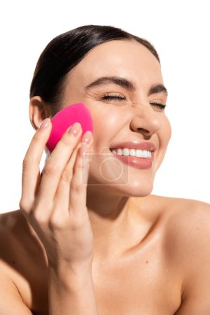 Lächelnde junge Frau schminkt Foundation mit rosa Schönheitsschwamm isoliert auf weiß 