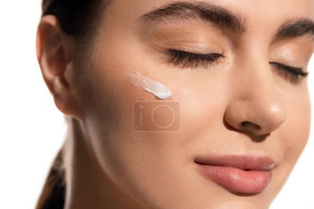 close up view of young woman with face cream on cheek isolated on white