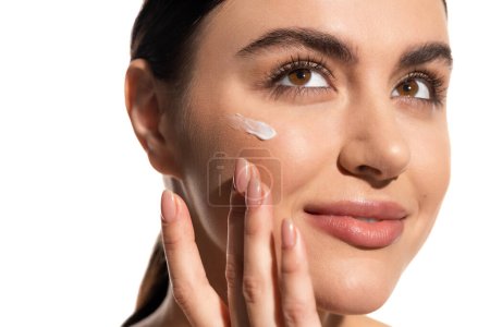 close up view of young woman with face cream on cheek touching face isolated on white