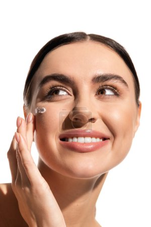 close up view of pleased young woman with face cream on cheek looking away isolated on white