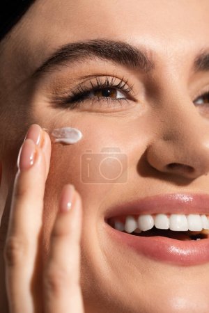 close up view of joyful young woman applying cream on cheek 