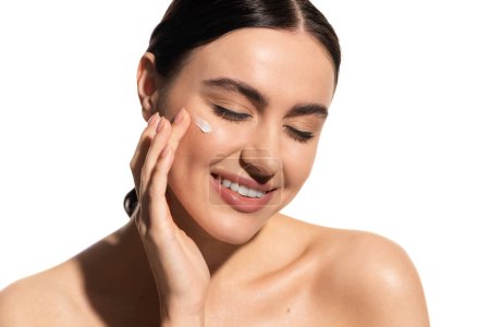 satisfied young woman applying moisturizing face cream on cheek isolated on white