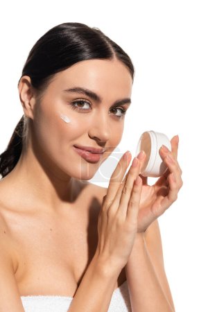 zufriedene junge Frau mit nackten Schultern hält Glas mit feuchtigkeitsspendender Gesichtscreme isoliert auf weiß 