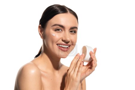 mujer joven positiva con hombros desnudos sosteniendo frasco con crema hidratante para la cara aislada en blanco 