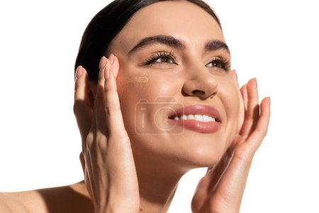 mujer joven feliz con la piel suave aplicando maquillaje fundación en la mejilla aislado en blanco 