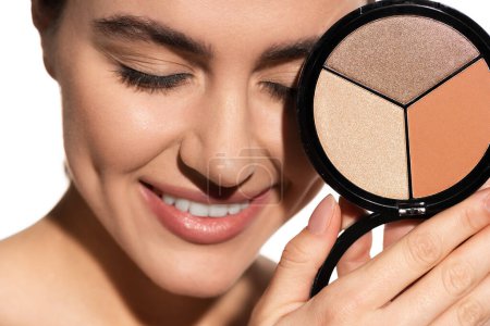 close up view of happy young woman holding bronzer and highlighter palette isolated on white 