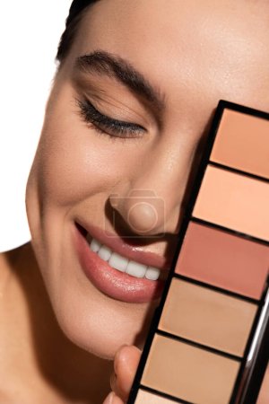 close up of positive young woman covering eye with eye shadow palette isolated on white 