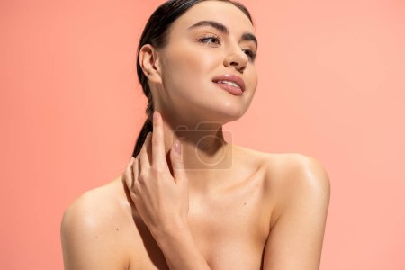 pleased woman with bare shoulders and flawless makeup touching soft skin isolated on pink 