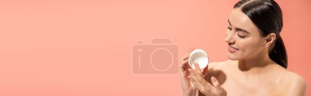Foto de Retrato de mujer complacida con hombros desnudos sosteniendo frasco con crema nutritiva aislado en rosa, bandera - Imagen libre de derechos