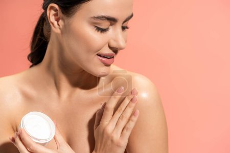 portrait of cheerful woman holding jar while applying nourishing cream on bare shoulder isolated on pink 