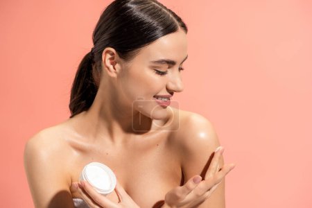 smiling woman holding jar and applying nourishing cream on bare shoulder isolated on pink 
