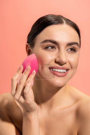 happy young woman applying makeup foundation with beauty sponge isolated on pink 