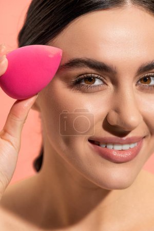 close up view of happy woman holding makeup sponge isolated on pink 