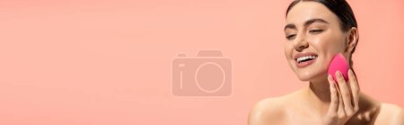 Téléchargez les photos : Femme heureuse appliquant fond de teint visage avec éponge beauté isolé sur rose, bannière - en image libre de droit