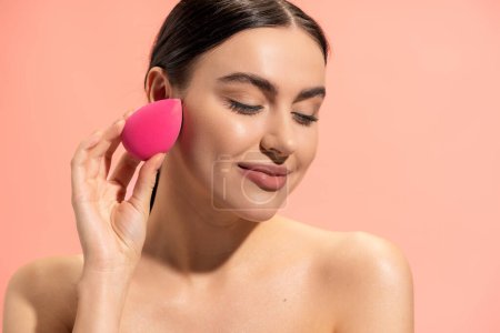 Téléchargez les photos : Femme heureuse appliquant fond de teint visage avec éponge de maquillage isolé sur rose - en image libre de droit