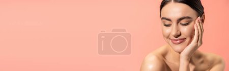 pleased woman with naked shoulders touching soft cheek while looking away isolated on pink, banner 