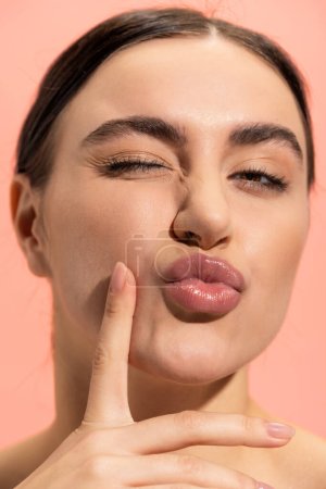 close up of young and brunette woman pouting lips isolated on pink 