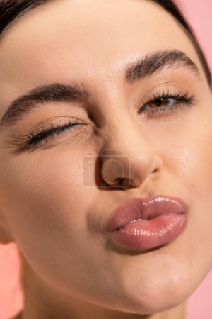 Nahaufnahme der jungen Frau schmollende Lippen und zwinkernde Augen isoliert auf rosa 