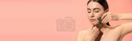 brünette Frau mit weicher Haut bei Gesichtsmassage mit Jadeschaber isoliert auf rosa, Banner 