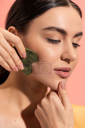 Téléchargez les photos : Vue rapprochée de la femme brune à la peau douce faisant massage du visage avec grattoir en jade isolé sur rose - en image libre de droit