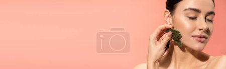 Téléchargez les photos : Brunette femme à la peau douce faisant massage du visage avec grattoir en jade isolé sur rose, bannière - en image libre de droit