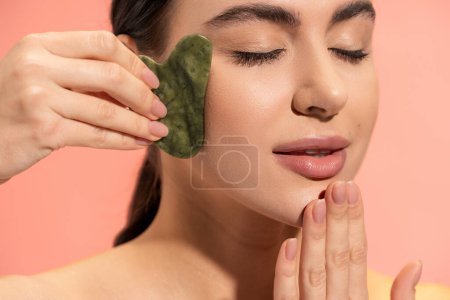 brunette woman with closed eyes doing massage with face scraper isolated on pink 