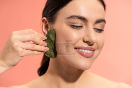 smiling woman with soft skin doing massage with face scraper isolated on pink 