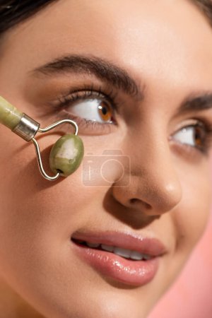 Foto de Close up view of smiling woman doing face massage with jade roller isolated on pink - Imagen libre de derechos
