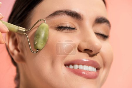 close up view of smiling woman with closed eyes doing face massage with jade roller isolated on pink  