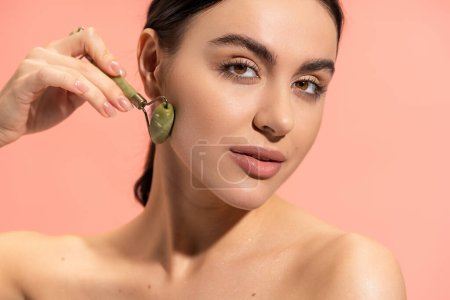young woman looking at camera while doing face massage with jade roller isolated on pink 
