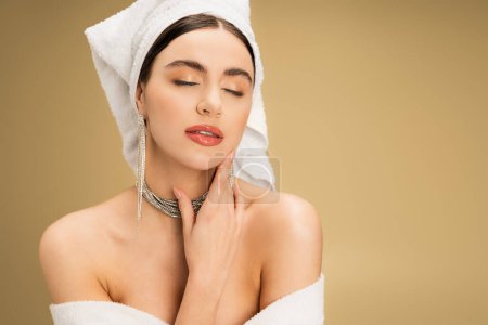 Téléchargez les photos : Élégante jeune femme avec maquillage et bijoux luxueux posant les yeux fermés sur fond beige - en image libre de droit