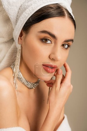 elegante mujer joven con maquillaje y joyas mirando a la cámara en gris 