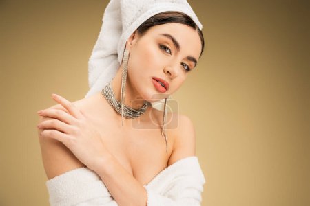 sensual woman with makeup and white towel on head touching bare shoulder on beige background 