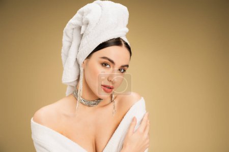 Foto de Charming woman in jewelry with white towel on head looking at camera on beige background - Imagen libre de derechos