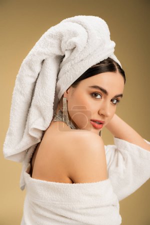 brünette Frau mit Make-up und Handtuch auf dem Kopf schaut in die Kamera auf beigem Hintergrund 