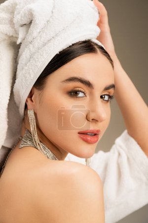 brünette Frau mit Make-up berühren weißes Handtuch auf dem Kopf auf grauem Hintergrund 